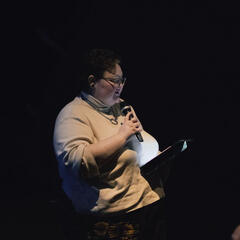 A photograph of Jenni, a person with a mousey brown mullet, clear acrylic glasses, presenting at the Accessify launch conference. Jenni is wearing a cream turtleneck jumper and black trousers with sunflowers on.